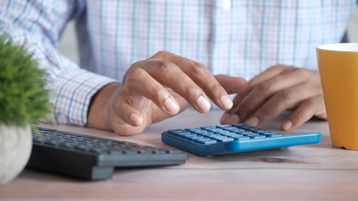 Dono de pequeno negócio de camisa xadrez fazendo contas numa calculadora azul sobre a mesa do escritório, ao lado do teclado e de uma xícara